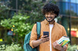 © Tetiana - Indian smiling young male student standing outside with backpack and books and using mobile phone