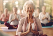 An elderly woman sits in the lotus position with her palms clasped while practicing meditation in a hall for group yoga and meditation classes.
