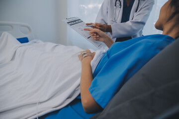 The senior woman specialist doctor with stethoscope checking up happy young Asian female patient lying in bed with receiving an intravenous saline drip in recovery room. Healthcare, medical insurance.