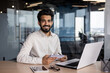 © Tetiana - Portrait of a young Indian successful man smiling and looking at the camera, sitting in the office at a desk with a laptop, holding a notebook