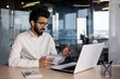 © Tetiana - Indian young male businessman working in office, sitting at desk holding notebook and talking on video call via laptop with clients and partners
