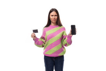 Wall Mural - young slender caucasian woman with straight black hair is dressed in a striped pink pullover holding a mock-up of a bank card and a smartphone. e-business concept