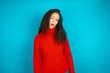 Beautiful teen girl wearing knitted red sweater over blue background winking looking at the camera with sexy expression, cheerful and happy face.