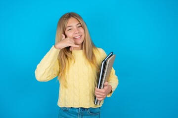 Wall Mural - Beautiful kid girl wearing yellow sweater holding notebook smiling doing phone gesture with hand and fingers like talking on the telephone. Communicating concepts.