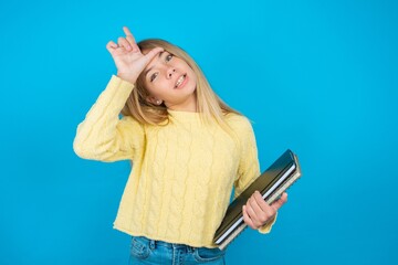 Wall Mural - Beautiful kid girl wearing yellow sweater holding notebook making fun of people with fingers on forehead doing loser gesture mocking and insulting.