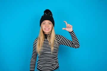 Wall Mural - Teen caucasian girl wearing striped sweater and woolly hat smiling and confident gesturing with hand doing small size sign with fingers looking and the camera. Measure concept