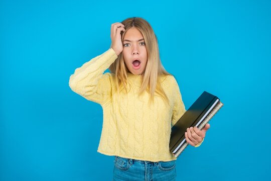 Embarrassed Beautiful kid girl wearing yellow sweater holding notebook with shocked expression, expresses great amazement, Puzzled model poses indoor