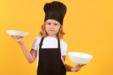 Wall Mural - Little cook with cooking plate. Child cooking, little chef prepares food. Kid boy in chefs hat and apron on yellow studio isolated background.