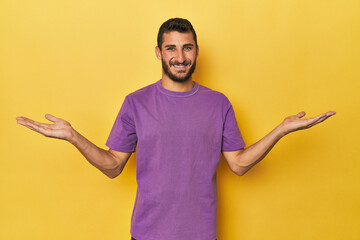 Young Hispanic man on yellow background makes scale with arms, feels happy and confident.