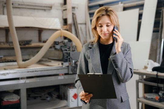 Portrait young woman in jacket use phone for call to client on background of wood workshop. Girl owner of workplace furniture carpenter with wood, small Business concept
