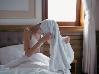 Young woman lying on bed with moisturizing beauty face mask and white towel on her head after shower, lifestyle home skincare, smile.