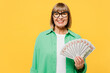 Elderly smiling fun blonde woman 50s year old wear green shirt glasses casual clothes hold in hand fan of cash money in dollar banknotes isolated on plain yellow background studio. Lifestyle concept.