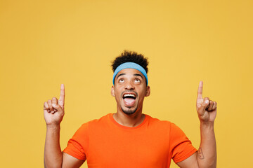 Wall Mural - Young fitness trainer instructor sporty man sportsman wear orange t-shirt point index finger overhead on area spend time in home gym isolated on plain yellow background. Workout sport fit abs concept.