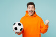 © ViDi Studio - Young happy man fan he wears orange hoody casual clothes do winner gesture cheer up support football sport team hold in hand soccer ball watch tv live stream isolated on plain blue color background.