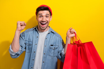 Wall Mural - Photo of cheerful lucky man dressed jeans shirt rising fist enjoying shopping isolated yellow color background