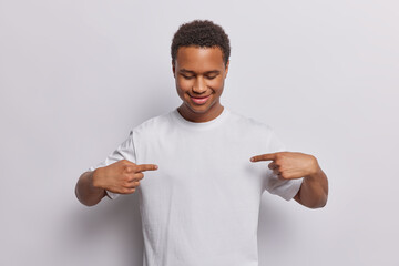 Wall Mural - Waist up shot of cheerful dark skinned man points index fingers at empty space of casual t shirt drawing attention to advertisement on it smiles gladfully isolated over white studio background