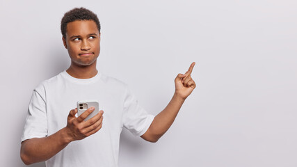 Wall Mural - Serious dark skinned man holds smartphone and points index finger at blank space presses lips wears casual tshirt shows place for your advertisement isolated over white background. Hmm look at this
