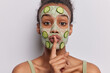 Indoor photo of young pretty European lady standing in centre isolated on white background wearing olive undershirt applying clay face mask and slices of cucumber making gesture to be silent