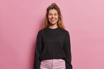 Poster - Horizontal shot of pretty young woman with long hair smiles broadly dressed in casual black sweater and trousers being in good mood expresses positive emotions isolated over pink backgground