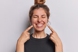 Fototapeta  - Photo of pleasant looking cheerful woman points index fingers at toothy smile shows wwell cared whie even teeth dressed in casual black t shirt isolated over whtie studio background feels happy