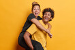 People positive emotions concept. Studio shot of young happy smiling standing Hindu male holding glad excited European female on back isolated in centre on yellow background wearing casual clothes