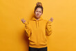 People emotions concept. Indoor photo of young confused European girl wearing yellow hoodie and black trousers standing in centre isolated spreading palms in hesitation not knowing what to do