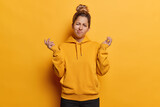 Fototapeta  - People emotions concept. Indoor photo of young confused European girl wearing yellow hoodie and black trousers standing in centre isolated spreading palms in hesitation not knowing what to do