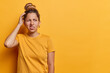 © WHstudio Leushin N - Waist up shot of European woman with hair bun scratches head and looks with bewilderment concentrated aside wears casual t shirt isolated over yellow background copy space for your promotional content