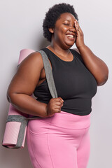 Wall Mural - Vertical shot of plump dark skinned woman makes face palm smiles positively as gains results in loosing weight dressed in sportswear carries fitness mat going to have pilates or cardio training.