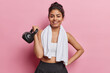 Horizontal shot of cheerful motivated Iranian woman lifts weight and smiles broadly poses with towel around neck dressed in activewear poses against pink background. Sport and exercising concept