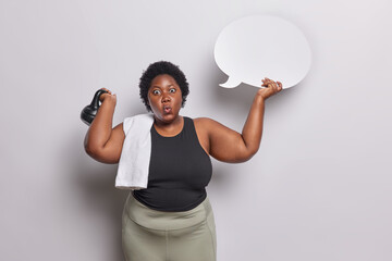 Wall Mural - Body positive dark skinned curly woman shows blank communication bubble for your advertisement and lifts weight has biceps workout session dressed in activewear isolated over white background