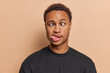 Studio close up of young funny African american man with short curly hair wearing black t shirt standing in centre isolated on beige background keeping hands down showing tongue making grimace