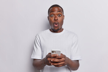 Wall Mural - Waist up shot of stunned dark skinned adult man holds mobile phone in hands shocked being bullied online reads unexpected message dressed in casual basic t shirt isolated over white background