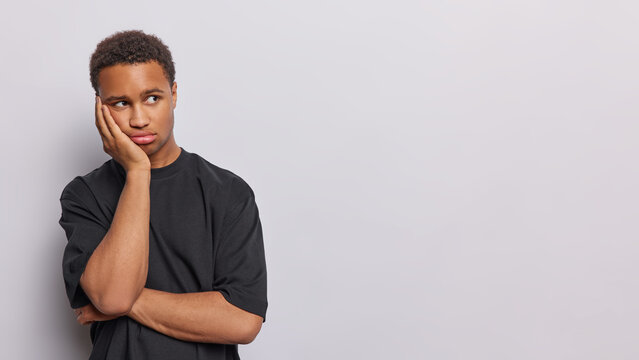 Pensive bored handsome African man keeps hand on cheek concentrated aside being deep in thoughts dressed in casual black t shirt isolated over white background mock up space for your advertisement