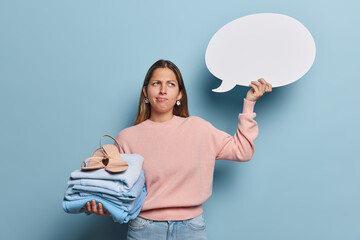 Wall Mural - Horizontal shot of thoughtful long haired woman holds folded clothes and shoes holds blank speech bubble for your promotion dressed casually isolated over blue background. Place your idea here