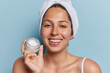 Beautiful positive woman with toothy smile hold jar of cream for skin treatment wears bath towel on head undergoes beauty procedures isolated over blue background. Wellbeing treatment concept