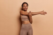 Sporty slim dark skinned woman stretches arms before workout dressed in cropped top and leggings has flat belly looks self assured at camera poses against brown background leads healthy lifestyle