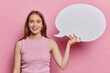 Waist up shot of cheerful long haired young woman dressed in casual t shirt smiles broadly suggests to write your promotional text on speech bubble poses against pink background. Hey great idea