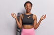 Slim dark skinned woman doing yoga and meditation dressed in cropped black top and leggings poes with fitness mat on shoulder isolated over white studio background. Healthy lifestyle concept