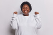 Waist up shot of glad overweight woman with curly hair waits announcement of results with hopeful cheerful expression smiles gladfully winks eye dressed in casual jumper isolated over white background