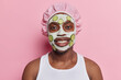 Horizontal shot of handsome pleased young African man wears bath hat and white t shirt applies beauty mask with cucumber slices for skin care isolated over pink background. Skin care procedures