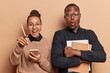 Horizontal shot of female and male groupmates pose with spiral notebooks and textbooks make notes react to something surprising point at camera isolated on brown background. People studying concept