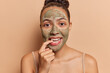 Horizontal shot of pretty Latin woman applies nourishing clay mask bites finger and looks directly at camera stands bare shouldered isolated on brown background. Beauty treatments and wellness concept