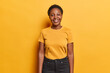 Indoor shot of good looking African with short curly keeps arms down smiles happily dressed in casual t shirt and black trousers looks directly at camera isolated over vivid yellow background