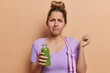 Horizontal shot of displeased European woman has sulking expression wears sportswear holds bottle of smoothie poses with skipping rope on shoulder goes in for sport regularly isolated on brown wall