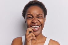 Studio Shot Of Dark Skinned Adult Woman With Curly Hair Bites Finger And Smirks Face Shows White Teeth Poses Against White Background Winks Eye Has Happy Expression. People And Emotions Concept