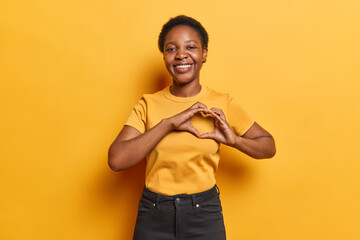 Horizontal shot of African woman shows heart love sign smiles gladfully dressed in casual t shirt and black trousers isolated over vivid yellow background. People and romantic feelings concept