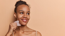 Cosmetic Natural Trend. Pretty Dark Skinned Woman With Dark Hair Gathered In Bun Smiles Tenderly Uses Gua Sha Quartz Stone For Massage Focused Aside Poses Against Brownn Background Copy Space