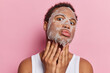 Skin care procedures. Dark skinned young African man applies beauty sheet mask on face for reducing fine lines and pores keeps lips rounded concentrated at camera isolated over pink background.