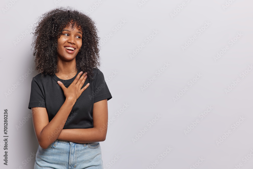 Horizontal shot of cheerful African woman with curly hair keeps hand on ...